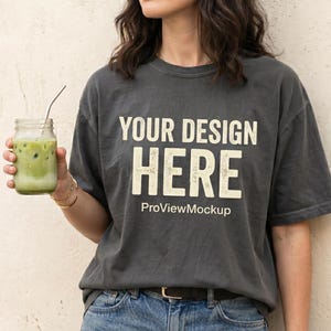 May include: Dark gray t-shirt with the text "YOUR DESIGN HERE" in a distressed white font. The shirt is paired with blue jeans and a gold watch. A glass jar with a green beverage and a metal straw is held in the left hand.