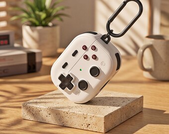 Könnte beinhalten: Eine weiße Schutzhülle für kabellose Ohrhörer, die wie ein Retro-Videospiel-Controller gestaltet ist. Sie verfügt über ein schwarzes Steuerkreuz, zwei schwarze Joysticks, vier mit A, B, X und Y beschriftete Tasten und einen schwarzen Karabinerhaken.
