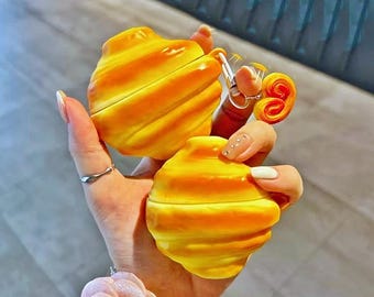Könnte beinhalten: Zwei gelb-orangefarbene Hüllen in Croissantform, möglicherweise für Ohrhörer, werden in einer Hand gehalten. Die Hüllen haben ein strukturiertes, geschichtetes Design. Ein kleiner Schlüsselanhänger mit einem Gebäck-Anhänger ist an einer Hülle befestigt. Der Hintergrund ist unscharf.