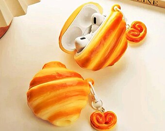 Könnte beinhalten: Eine ausgefallene AirPod-Hülle in Form eines Croissants, mit passendem Anhänger. Die Hülle ist geöffnet und zeigt weiße Ohrhörer. Hülle und Anhänger sind in Gelb- und Brauntönen gehalten und ähneln Backwaren.