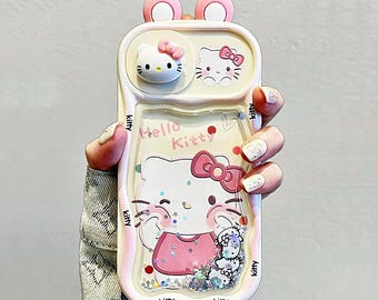 Könnte beinhalten: Eine hellrosa Handyhülle mit einem Hello Kitty-Design. Die Hülle hat eine 3D-Hello Kitty-Figur, die Worte "Hello Kitty" und einen glitzernden, flüssigkeitsgefüllten Bereich. Die Hülle hat auch rosa, ohrenförmige Akzente oben.