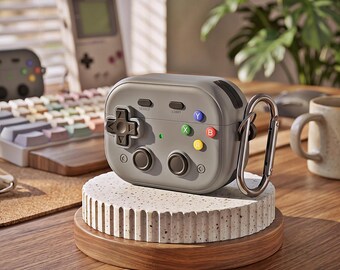 Könnte beinhalten: Eine matte graue Schutzhülle für kabellose Ohrhörer, die wie ein Retro-Videospiel-Controller gestaltet ist. Sie verfügt über ein schwarzes Steuerkreuz, zwei Analogsticks, farbige Aktionstasten mit Y, X, B, A Beschriftung und einen Metallkarabiner.