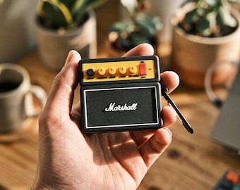 Könnte beinhalten: Eine schwarze Schutzhülle für kabellose Ohrhörer, die wie ein Miniatur-Gitarrenverstärker gestaltet ist. Die Hülle verfügt über ein gelbes Bedienfeld mit Reglern für Input, Volume, Gain, Tone, Reverb sowie einen roten Ein-Aus-Schalter. Auf der Vorderseite befindet sich das weiße Marshall-Logo.