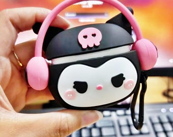 Könnte beinhalten: Schwarzes und weißes Cartoon-AirPods-Case mit rosa Kopfhörern und einem Totenkopf-Emblem. Das Case hat ein weißes Gesicht, schwarze Augen und rosa Wangen. Ein schwarzer Karabinerhaken ist angebracht.