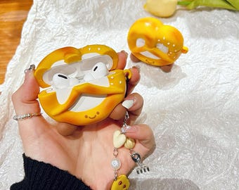 Könnte beinhalten: Eine gelbe, brezelförmige AirPod-Hülle, die geöffnet ist, um weiße Ohrhörer freizulegen. Die Hülle hat ein lächelndes Gesicht und einen Anhänger mit einem Keks und herzförmigen Perlen. Eine weitere brezelförmige Hülle ist im Hintergrund sichtbar.