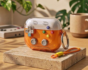 Könnte beinhalten: Eine durchscheinende orangefarbene Schutzhülle für kabellose Ohrhörer, die wie eine Retro-Handheld-Spielkonsole gestaltet ist. Sie verfügt über einen weißen oberen Bereich, ein graues Steuerkreuz, vier farbige Aktionstasten mit der Beschriftung A, B, X, Y sowie zwei graue Joysticks. Ein Metallring und eine orangefarbene Handschlaufe sind befestigt.