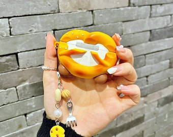 Könnte beinhalten: Eine gelb-orangefarbene Brezel-förmige Hülle für kabellose Ohrhörer. Die Hülle hat einen kleinen Anhänger mit Perlen, einer Miniatur-Gabel und einem gelben Keks-Anhänger. Die Hülle wird in einer Hand vor einer grauen Backsteinmauer gehalten.