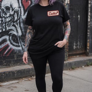 May include: A black short-sleeved t-shirt featuring a rectangular patch on the left chest with a red border and the name Debbie written in a cursive script font, accompanied by a small red coffee cup icon above the name.