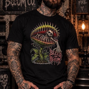 May include: A black short-sleeve t-shirt featuring a graphic print of a skeletal figure inside a large, spiky, green and yellow carnivorous plant. The text 'BLEED AND BLOOM' is printed in a stylized red font below the plant illustration.