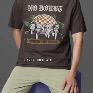 May include: Dark chocolate-colored t-shirt featuring the band No Doubt with the text "No Doubt" in a vintage font. The shirt also has the text "Sphere Las Vegas" and a checkered sphere graphic.