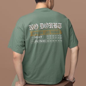 May include: Sage green t-shirt with the text "NO DOUBT" in a vintage font. Below the text is a calendar grid for May and June 2020. The person is sitting on a brown cube.