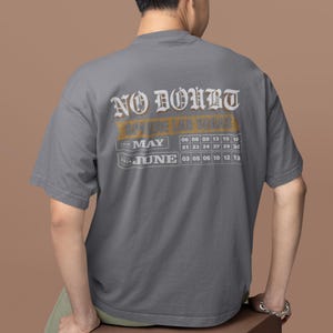 May include: Gray t-shirt with the text "NO DOUBT" in a vintage font, with "STILL IN LAS VEGAS" below. The shirt includes a calendar for May and June 2020. The person is sitting on a brown cube.