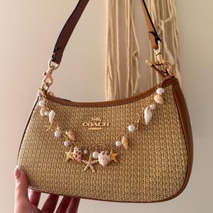 Peut inclure: Un sac à main Coach avec un corps en paille tressée et des bordures en cuir marron. Le sac est orné d'une chaîne décorative avec des coquillages, des perles et des breloques en forme d'étoiles de mer. Il a une anse en cuir marron et des accessoires dorés.