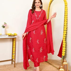 May include: A vibrant red long-sleeved kurta set featuring delicate floral embroidery in pink and white thread, accented with small silver beads. The ensemble includes a matching red dupatta draped over the arms and coordinating red trousers.