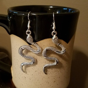 May include: A pair of silver snake earrings with a textured finish. The earrings are hanging from a mug.