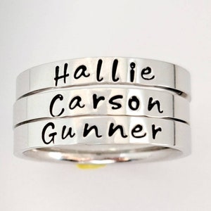 May include: Three silver rings stacked on top of each other. The rings are engraved with the names "Hallie", "Carson", and "Gunner".