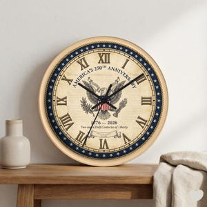 May include: A round wall clock with a cream-colored face and a beige frame. The clock features Roman numerals and the text "America's 250th Anniversary" with an eagle emblem. The clock's face also includes the years 1776-2026.