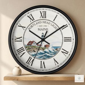 Op de afbeelding: Ronde wandklok met een zwart frame en een witte wijzerplaat. De wijzerplaat toont Romeinse cijfers en de tekst "Portland Head Light - Est. 1791 - Maine." De klok toont een vuurtoren.