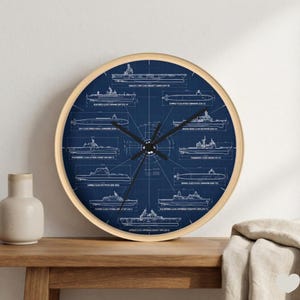 May include: A circular wall clock with a light wood frame. The dark blue clock face features a blueprint design of various ships with text. Black clock hands show the time.