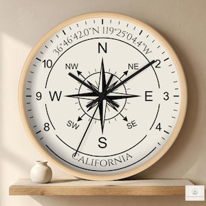 Op de afbeelding: Ronde wandklok met een licht houten frame en een crèmekleurige wijzerplaat. De klok heeft een kompasroosontwerp met zwarte accenten, het woord "CALIFORNIA" en coördinaten. De wijzers zijn zwart.
