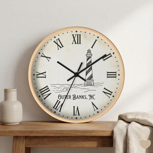 Könnte beinhalten: Runde Wanduhr mit hellem Holzrahmen und cremefarbenem Zifferblatt. Die Uhr hat schwarze römische Ziffern und Zeiger. In der Mitte befindet sich eine schwarz-weiße Illustration eines Leuchtturms mit dem Text "Outer Banks, NC" darunter.