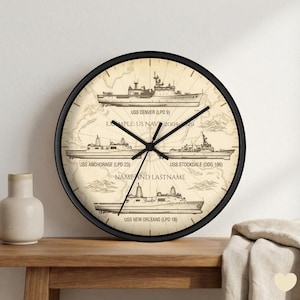 May include: A round wall clock with a black frame. The clock face features a vintage map design with illustrations of naval ships and text including "USS DENVER", "USS ANCHORAGE", "USS STOCKDALE", and "USS NEW ORLEANS". The clock hands are black.