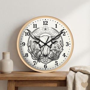 May include: A round wall clock with a light wood frame. The clock face features a black and white illustration of a bull skull, cacti, and a starburst design. Black numbers mark the hours.