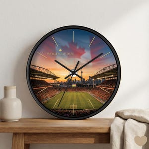 Puede incluir: Un reloj de pared redondo con marco negro. La esfera del reloj muestra una escena de estadio con cielo de atardecer, la bandera canadiense y el texto "WORLD CUP 2026". El reloj está sobre una superficie de madera.