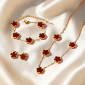 Può includere: Un set di gioielli color oro con collana, bracciale e orecchini. Ogni pezzo presenta un design a trifoglio con centri in smalto rosso. I gioielli sono esposti su una superficie bianca e setosa.