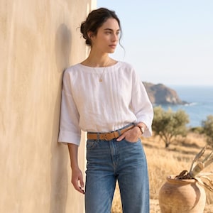 May include: A white linen blouse with three-quarter sleeves and vertical pleat detailing paired with high-waisted blue denim jeans, a tan leather belt, and tan leather loafers. The outfit is completed with a simple gold pendant necklace.
