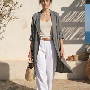 Peut inclure: Un long manteau gris ouvert sur le devant avec des manches trois-quarts, porté sur un débardeur beige à encolure dégagée et un pantalon blanc à jambes larges. Une ceinture en cuir beige à boucle dorée marque la taille, avec un sac en paille.