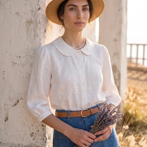 May include: A white button-down shirt with a rounded Peter Pan collar and three-quarter length sleeves, paired with a blue denim skirt, a tan leather belt, a straw fedora hat, and a simple white beaded necklace, holding a bundle of dried lavender.