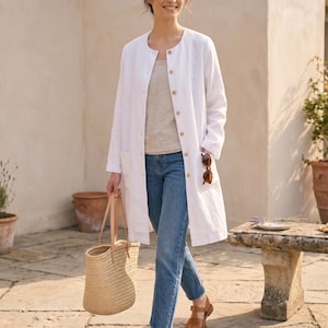 May include: A long-sleeved white linen coat with a round neckline and a front button closure, worn over a beige top and blue denim jeans with a frayed hem. The outfit includes brown leather huarache sandals and a woven straw tote bag with leather handles.