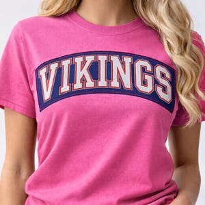 May include: A pink t-shirt with the word "VIKINGS" in white, outlined in red, on a navy blue arched background. The shirt is short-sleeved and has a crew neck.
