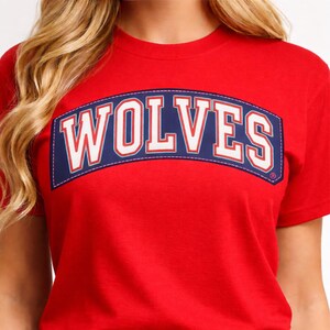 Puede incluir: Una camiseta roja con la palabra "WOLVES" en letras blancas sobre un parche arqueado azul marino. El parche tiene un borde blanco con costuras rojas. La camiseta es de manga corta.