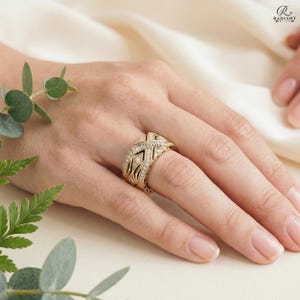 A woman's hand wearing a sparkling 14K gold geometric diamond Oura ring cover on her index finger. The hand rests against elegantly folded ivory fabric and fresh green leaves.