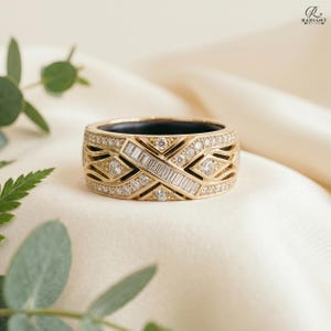 Close-up of a 14K gold Oura Ring Gen 4 cover featuring a geometric criss-cross design with baguette and round lab-grown diamonds, resting on soft cream fabric with green botanical leaves.