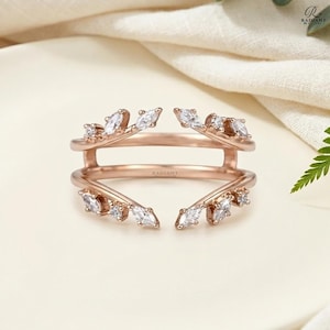 Peut inclure: Garde de bague en or rose ornée de pierres blanches de taille marquise et ronde. La bague présente un design délicat d'inspiration florale avec le mot "RADIANT" gravé sur le corps. La bague est posée sur un fond neutre et doux.