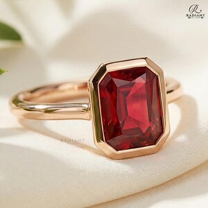 Könnte beinhalten: Ein roségoldener Ring mit einem großen, rechteckigen, tiefroten Edelstein. Der Edelstein ist in einer Lünettenfassung gefasst, und das Band ist glatt und poliert. Der Ring wird auf einer weißen, strukturierten Oberfläche präsentiert.