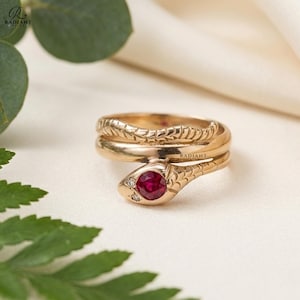May include: A gold-colored snake ring with a ruby gemstone and small diamond accents. The ring is coiled, with the snake's head at the front. The ring is on a white surface with green foliage in the foreground.