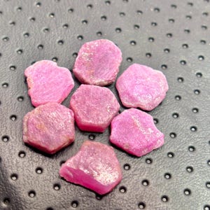 May include: A collection of seven rough-cut ruby gemstones. The rubies display a vibrant pinkish-red hue, with irregular shapes and a natural, unpolished texture. The gemstones are arranged on a dark, textured surface.
