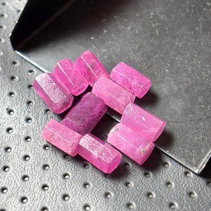 May include: A collection of faceted, pink-red gemstones. The gemstones are rectangular with beveled edges, and appear to be raw or unpolished. The gems are scattered on a dark, textured surface, with a metal tray in the background.