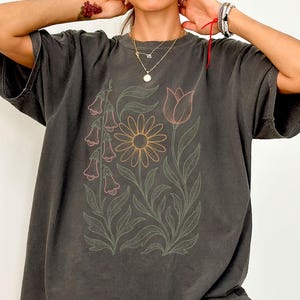 May include: Dark gray t-shirt featuring a floral design. The graphic includes a daisy, tulip, and bell-shaped flowers, outlined in pink and orange, with green leaves and stems. The shirt has short sleeves and a relaxed fit.