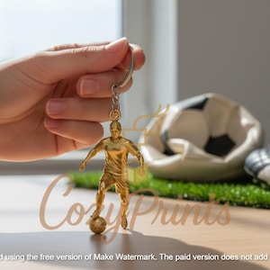 Peut inclure: Un porte-clés doré représentant un joueur de football. La figurine est posée avec un ballon de football à ses pieds. Un ballon de football dégonflé et une paire de chaussures de football noires et blanches sont en arrière-plan. Le mot "CozyPrints" est visible.