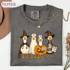 May include: A charcoal gray t-shirt featuring a graphic of four ducks dressed in Halloween costumes. One duck wears a witch hat and cape with a stethoscope, one is a ghost, one is inside a pumpkin, and one is a skeleton wearing a witch hat.