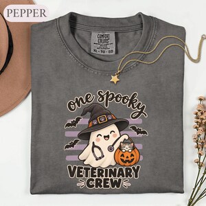 May include: A charcoal gray t-shirt featuring a graphic of a ghost wearing a witch hat and a stethoscope, holding a small pumpkin with a kitten inside. The text reads one spooky VETERINARY CREW. The shirt has a Comfort Colors label in the neck.