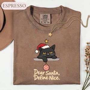 May include: An espresso-colored t-shirt featuring a graphic of a black cat wearing a red Santa hat, resting on a ledge while holding a pink Christmas ornament. Below the cat, the text reads, "Dear Santa, Define Nice." The shirt has a Comfort Colors label.