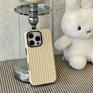 May include: A yellow and white striped phone case. The case has a black camera lens cutout and black side buttons. A plush white bunny is in the background, along with a silver candle holder.