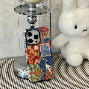 May include: A colorful phone case with a patchwork design featuring floral, cat, and sun illustrations. The case has a black camera cutout and a white border. The case is on a silver stand with a white wooden background.