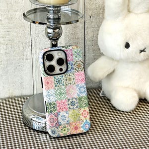 May include: A smartphone case featuring a multi-coloured tile pattern with various floral and geometric designs in shades of pink, green, blue, and yellow. The case has a black camera cutout frame and covers the back of a mobile device.